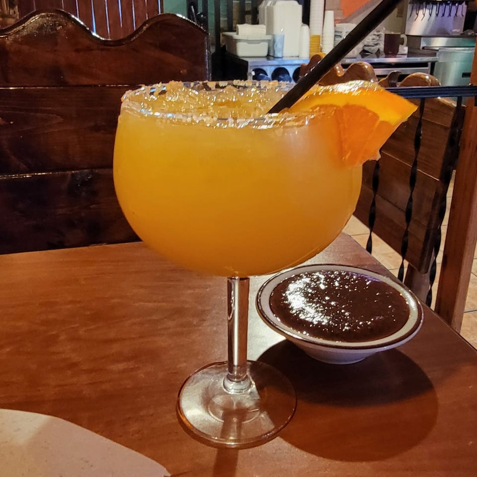 A large La Casona margarita with a salted rim and orange slice, next to a bowl of salsa.