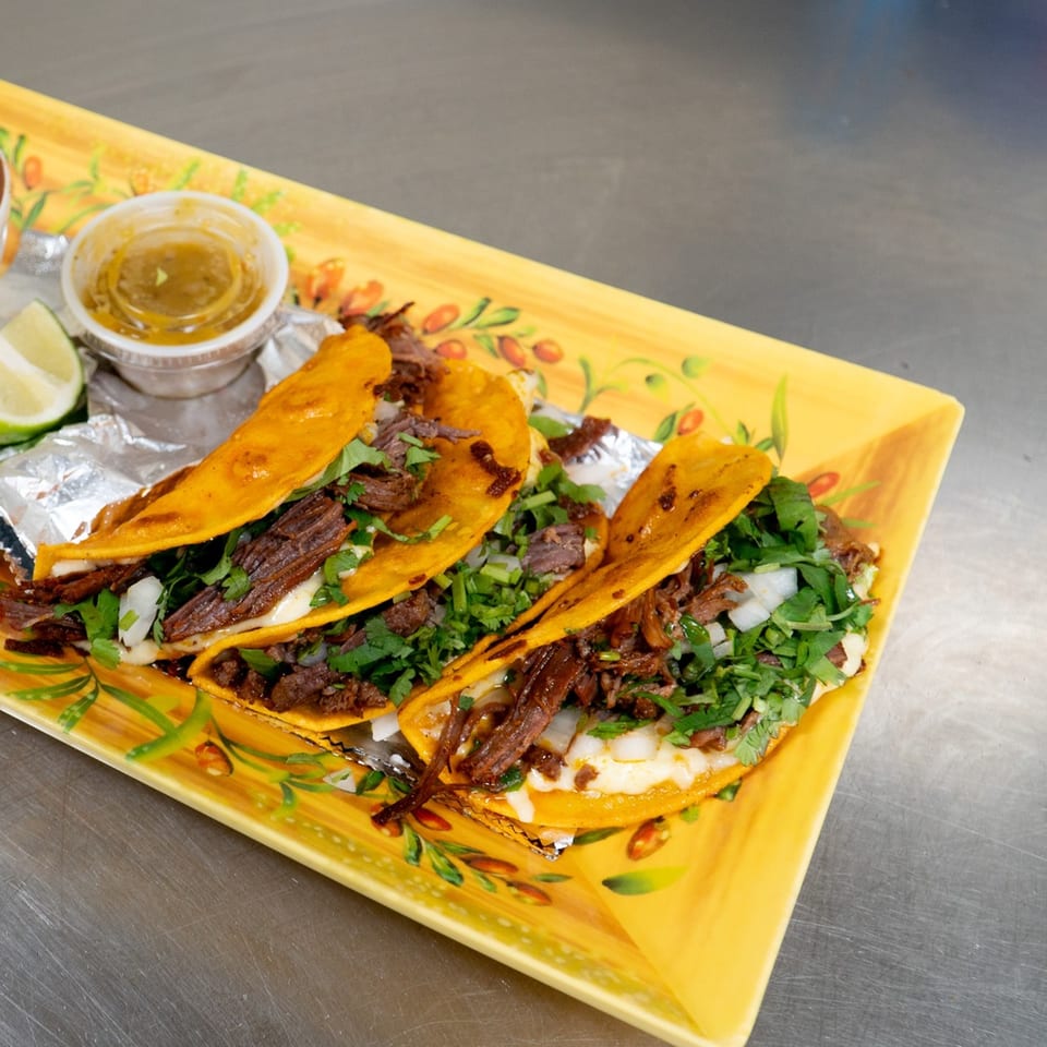 Three La Casona Mexican Restaurant birria tacos with cilantro, onions, and lime on a yellow plate.